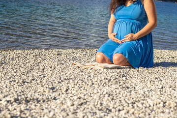 Fototapeta na wymiar woman big advanced pregnancy walking along lake side on pebbles beach.third last trimester female in blue dress mountain landscape clear turquoise water.baby awaiting mom holding caressing belly
