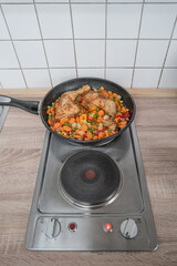 Electric stove in the kitchen with a frying pan cooking chicken legs and vegetables, with copy space background. Concept food and economic crisis
