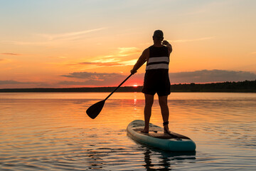 A man in shorts stands on a sapboard with a paddle in the evening at sunset in a lake in the sun glare.