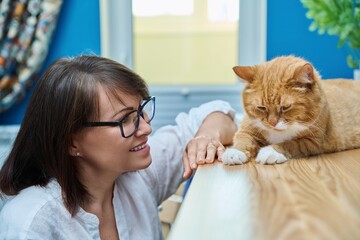 Middle aged woman talking with ginger pet cat, home interior background
