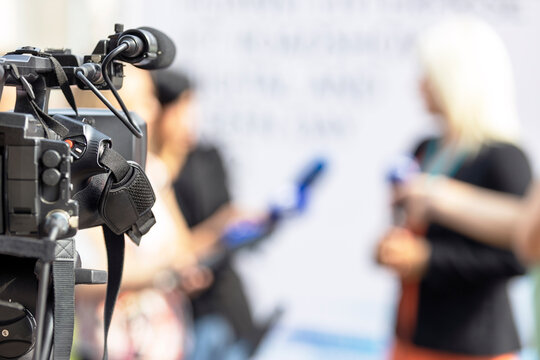 Video camera in focus, blurred journalists at news conference holding microphones making media interview with female politician or business woman