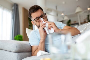 Sick sad man with glasses sits on couch at home suffers from runny nose flu disease coronavirus...