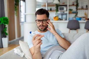 Man feeling sick lying in the bed and looking the thermometer. Sick man lying on sofa checking his...