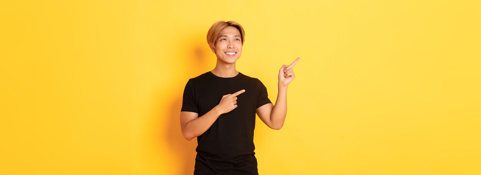Portrait of handsome korean blond guy, pointing fingers upper left corner and looking satisfied with pleased smile, yellow background