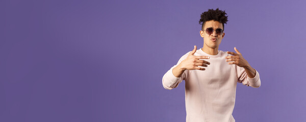 Portrait of sassy and carefree young hipster guy with dreads, wearing sunglasses dancing and gesturing while singing during rap battle, dance hip-hop on party, standing purple background