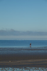 A lone fisherman at the seaside on the beach. A solitary fisherman seen from behind, feet in the water, holds his fishing rod alone in front of the ocean and the islands in the distance.