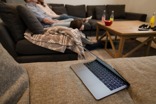 Laptop On Sofa With Family Watching TV On Sofa In Background