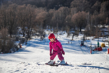 Fototapeta na wymiar Child girl skier standing on the ski slope of a snowy mountain in sunny winter day preparing for a downhill skiing trip on training