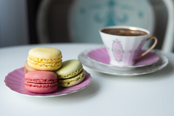 Mix of appetizing macarons and delicious coffee in porcelain cup on white table decorated with blurred flowers. Dry flowers. Greeting card.
