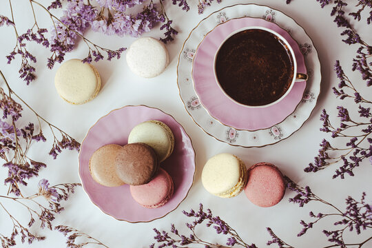 Top view of appetizing macarons and black coffee in porcelain cup on white table decorated with blurred flowers. Sweet concept. Flat lay. Excellent image for dessert banners and advertisements.