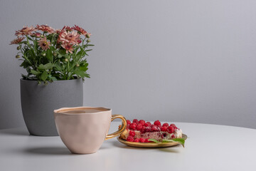 Delicious piece of cake with fresh raspberries and luxurious cup of coffee next to cake on white table. Beautiful pink chrysanthemums in grey pot in background. Light grey background. Place your text.