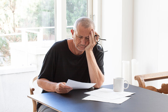 Frustrated man reading paper documents while sitting at table with glasses in hand holding his head.