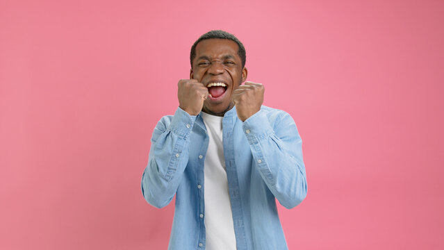 Happy Smiling 20 Year Old African American In Denim Shirt And White T-shirt, Makes Winner Gesture, Says Yes, Guy Is Emotionally Happy To Win Or Winning Victory Isolated On Pink Background In Studio.