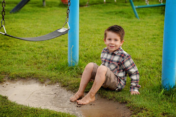 Niño feliz sentado mojando sus pies en un charco de agua con lodo y tierra disfrutando y jugando emocionado en el parque al aire libre en un hermoso día  - obrazy, fototapety, plakaty
