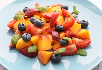 Fruit salad in a blue plate on a white background. Sliced strawberries, peaches and blueberries with mint leaves. Free space for text.