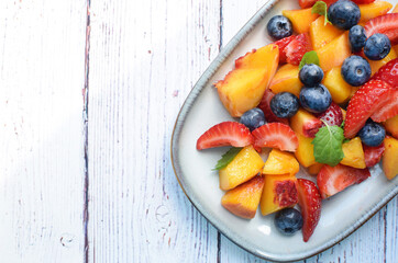 Fruit salad in a rectangular plate on a white wooden background. Sliced strawberries, peaches and blueberries with mint leaves.