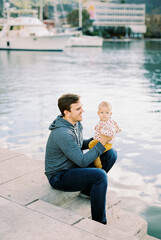 Dad sits on the pier with a baby on his lap against the backdrop of yachts
