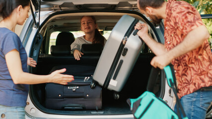 Parents loading voyage luggage inside car trunk while unpatient daughter hurrying them. Casual family going on summer holiday citybreak while loading baggage and trolleys inside vehicle. © DC Studio
