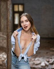 Stylish young woman with flowing long brown hair in knotted blue shirt posing near steel mesh fencing inside destroyed deserted building..