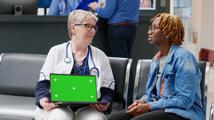 Senior doctor showing laptop with greenscreen template to patient sitting in waiting area lobby. Medic and person looking at blank copyspace with chroma key display and isolated mockup.