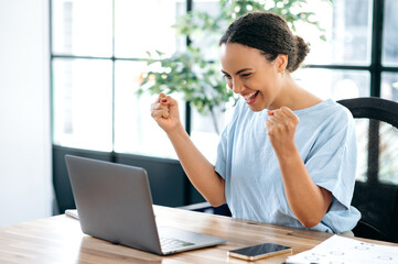 Joyful amazed happy brazilian or hispanic young business woman, sitting at a desk with a laptop in modern office, rejoicing in success, big profit, celebrate deal, gesturing with fists, smiling