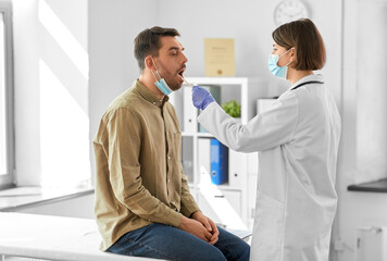 Obraz premium medicine, healthcare and people concept - female doctor in mask with tongue depressor checking sore throat of man patient at hospital