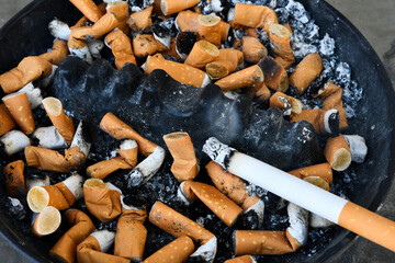 A close up image of a dirty unhealthy ashtray with a burning cigarette. 
