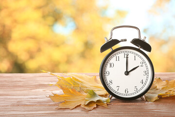 Alarm clock with autumn leaves on wooden table outdoors