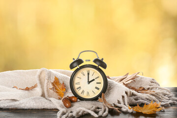 Alarm clock with knitted plaid and autumn leaves on wooden table outdoors