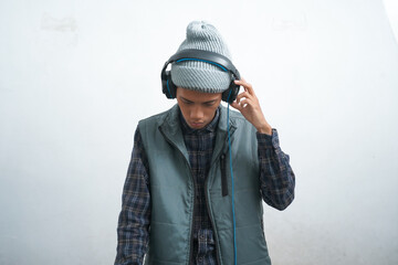 asian young man wearing dark blue vest and beanie wearing headphones, isolated on white background focused on listening to music via smartphone