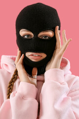 Portrait of young woman in balaclava touching face on pink background