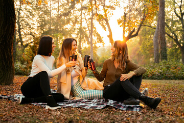 Group of three female friends smiling, sitting on picnic blanket in the park and drinking coffee,...