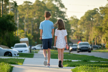 Rear view of two young teenage children, girl and boy, brother and sister walking together on rural street on bright sunny day. Vacation time concept