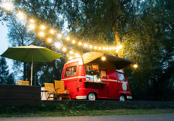 Empty small Street Food Track with an illuminations standing in the deserted night park. Near food trailer are folding tables and chairs - obrazy, fototapety, plakaty