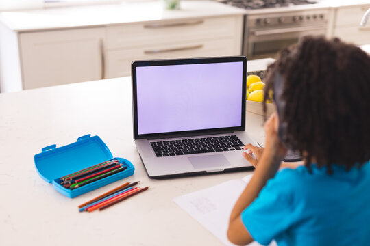 Rear view of hispanic boy using laptop for online classes while sitting at dining table - Powered by Adobe