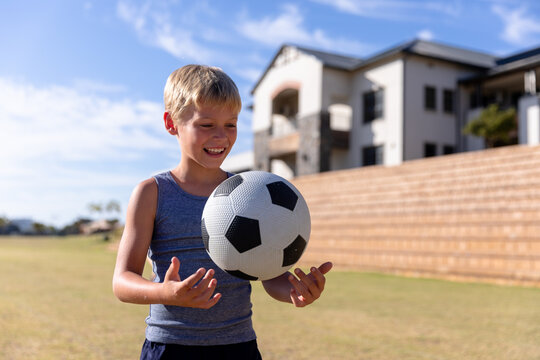 Smiling caucasian elementary schoolboy playing with soccer ball on field against school building