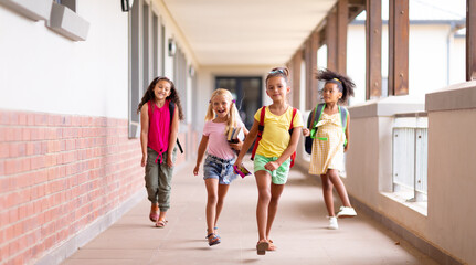 Full length of happy multiracial elementary schoolgirls walking in corridor