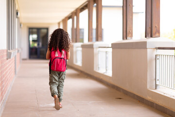 Rear view full length of biracial elementary schoolgirl with backpack walking in corridor at school