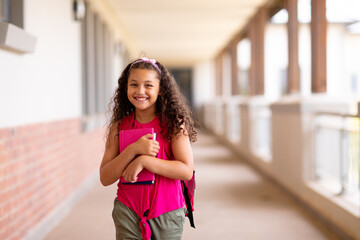 Portrait of smiling cute biracial girl holding books while standing in corridor at school