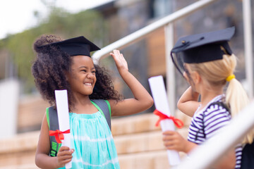 Cheerful multiracial elementary schoolgirls with mortarboards and degree doing high-five on steps