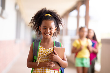 Portrait of smiling biracial elementary schoolgirl holding books while standing in corridor