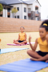 Naklejka premium Multiracial elementary schoolgirls meditating while sitting on yoga mat against school