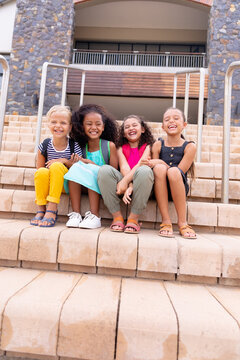 Full length of multiracial cheerful elementary schoolgirls sitting on steps against school building