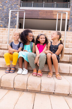 Full length of multiracial smiling elementary schoolgirls sitting on steps against school building