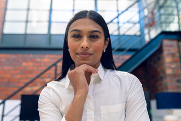 Portrait of smiling biracial young businesswoman with hand on chin sitting in office - Powered by Adobe