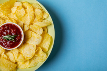 Red sauce in bowl amidst potato chips served in plate on blue background with copy space