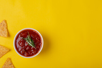 Red sauce with rosemary in bowl by nacho chips on yellow background with copy space