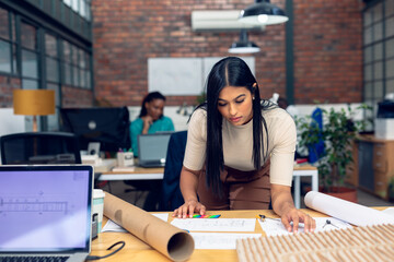 Biracial young female architect working over blueprint by building model at desk in office