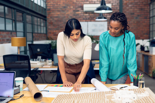 Biracial and african american young female architects discussing over blueprint at desk in office