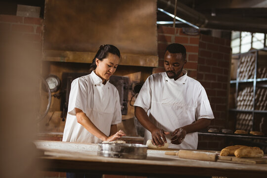 Asian mid adult female baker showing dough kneading to african american mid adult male coworker